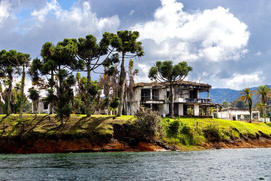 Abandoned house of Pablo Escobar stands on grassy cliff edge. Large trees frame the crumbling structure. Water laps at the shoreline below. Sky shows white clouds and blue patches
