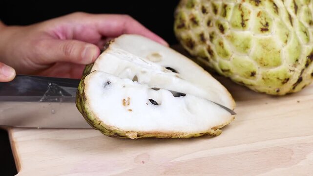A close up shot of a person using a sharp knife to slice a fresh sugar apple or custard apple on a wooden cutting board.