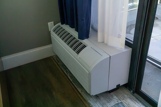 Modern PTAC (Packaged Terminal Air Conditioner) unit providing essential climate control in a comfortable room. Located by a window with blue and sheer curtains, a dark wall, and elegant wood