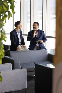 Two businessmen met in office lounge with digital tablet, discuss information during workday. Ideas and business solutions sharing, collaborative project review, communication, professional discussion