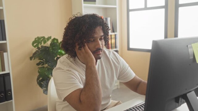 Man typing on computer with hand on face at desk in building, seated by monitor and plant, wearing white tshirt and showing weary posture; fatigue focus concentration thought.