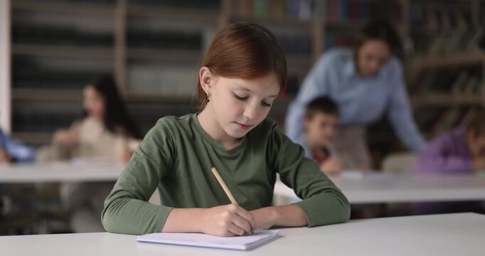 Smart schoolgirl child studying sitting at school desk in classroom, writing exercise in copybook, take knowledge test, making assignment during class. Primary school education, skill development