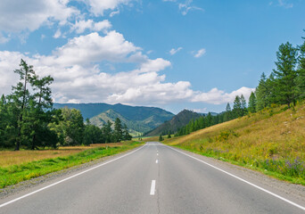 Fototapeta premium Long paved road extending through green hills and forests toward distant mountains under a beautiful summer sky