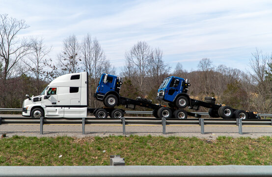 Long-haul white semi-truck transporting two new blue Mack cab-over truck chassis frames on a highway, illustrating heavy freight logistics and commercial vehicle delivery.