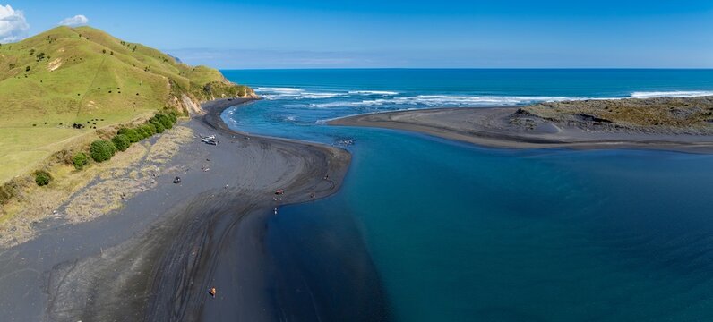 Aerial view of people enjoying a sunny day at the black sand beach and river mouth in Marokopa, New Zealand. Tourists and locals relax and swim in the water.