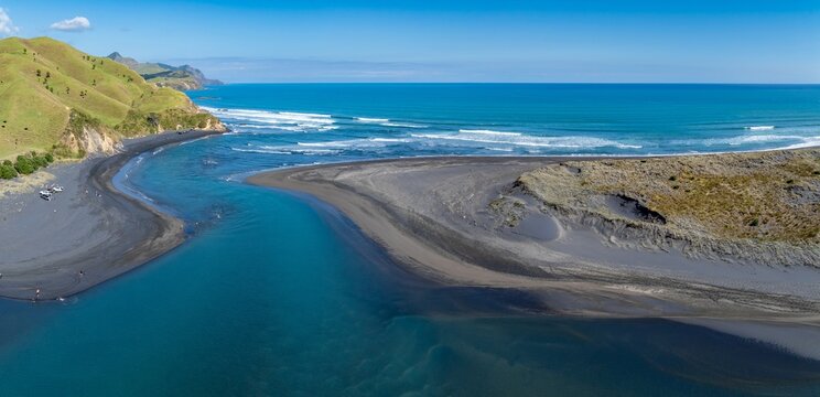 Aerial view of people enjoying a sunny day at the black sand beach and river mouth in Marokopa, New Zealand. Tourists and locals relax and swim in the water.