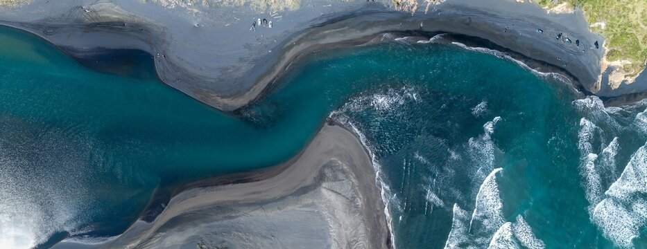 Aerial view of people enjoying the black sand beach and turquoise river mouth at Marokopa in New Zealand. Vehicles parked nearby for easy access.