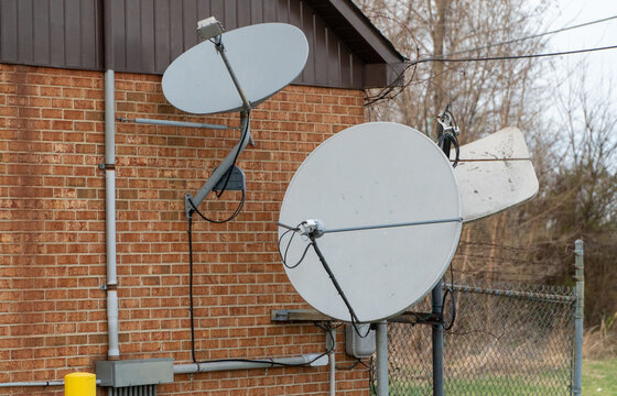 Two large gray satellite dishes mounted on a weathered red brick wall with brown metal siding, showcasing outdated communication technology, wires, and conduits. A chain-link fence with barbed wire