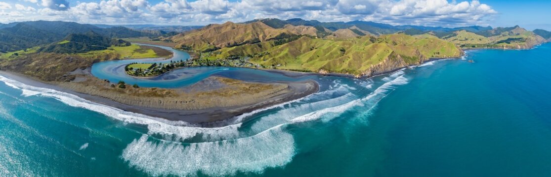 Aerial view of the scenic coastline of Marokopa, New Zealand. The turquoise ocean meets the black sand beach, with rolling green hills in the background. A small town sits by the river mouth.