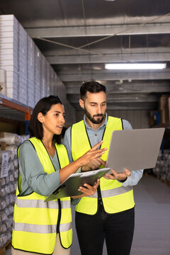 Warehouse employees standing next to storage racks, using laptop, checking inventory records, updating storehouse system. Logistics workflow, supply chain management, distribution center operations