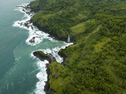 Aerial view of Banyutibo Water Fall cascading into the turquoise sea from rocky cliffs surrounded by lush terraced hills Girisubo, Special Region of Yogyakarta, Indonesia.