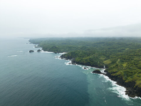 Aerial view of Banyutibo Water Fall cascading into the ocean from lush green cliffs under a misty sky Girisubo, Special Region of Yogyakarta, Indonesia.