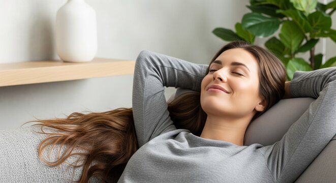 Serene brunette woman relaxing on sofa with hands behind head eyes closed in peaceful contentment wellness concept calm pose in gray clothing