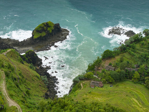 Aerial view of Banyu Tibo Waterfall cascading onto a rocky beach with turquoise ocean waves, lush green cliffs, and small coastal dwellings in Girisubo, Special Region of Yogyakarta, Indonesia.