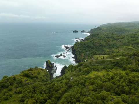Aerial view of Banyutibo Water Fall cascading onto the beach surrounded by lush green cliffs and terraced hills Girisubo, Special Region of Yogyakarta, Indonesia.