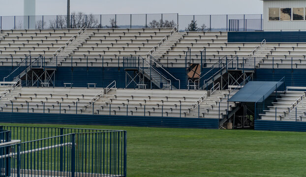 Large, empty stadium with rows of white and blue bleachers overlooking a vibrant green artificial turf field, ideal for athletic event themes.