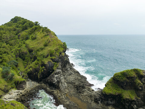 Aerial view of Banyutibo Water Fall cascading down a lush green cliff into the turquoise sea with crashing waves against the rocky coastline in Girisubo, Special Region of Yog, Indonesia.