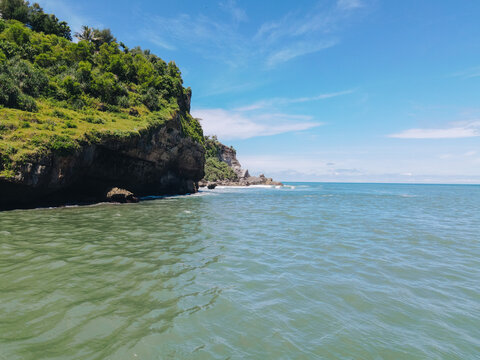 Aerial view of Baron Beach featuring rocky cliffs covered in lush vegetation and turquoise sea water under a clear blue sky Tanjungsari, Special Region of Yogyakarta, Indonesia.