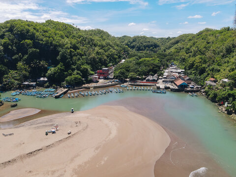 Aerial view of Baron Beach with numerous blue fishing boats moored along the river mouth and lush green hills Tanjungsari, Special Region of Yogyakarta, Indonesia.