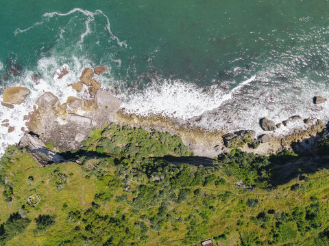 Aerial view of Baron Beach featuring rocky cliffs, crashing white waves, and lush green vegetation in Tanjungsari, Special Region of Yogyakarta, Indonesia.