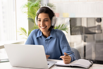 Naklejka premium Smiling businesswoman in headphones working or studying at laptop in office, engaged in online meeting or virtual training session, gains new skills, e-learns at workplace. Professional development