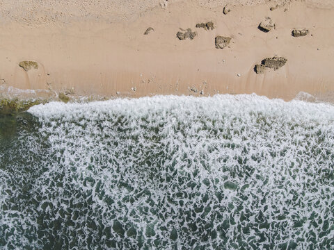 Aerial view of Baron Beach with white waves crashing on the sandy shore and scattered rocks in Tanjungsari, Special Region of Yogyakarta, Indonesia.