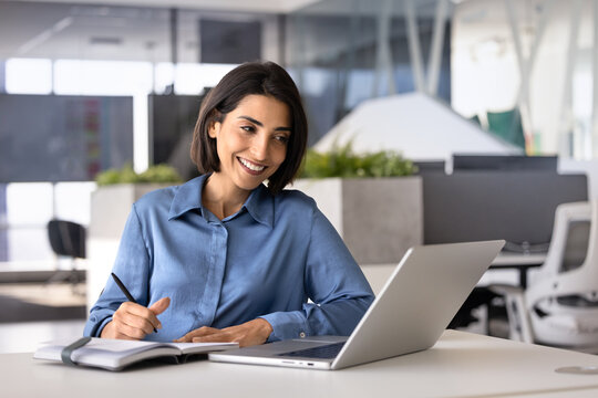 Smiling office employee studying at workplace desk watching educational content on laptop, engage in online meeting or webinar, make notes, writing information in notebook, improve professional skills