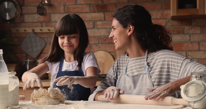 Caring mother with daughter flattening and kneading dough together in domestic kitchen. Family cooking, culinary hobby, learning activity, parent sharing knowledge with child, household chores at home