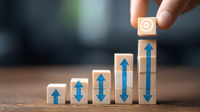 Hand placing a wooden block with target symbol on an ascending stack with blue up-down arrows, representing goal achievement and progress.