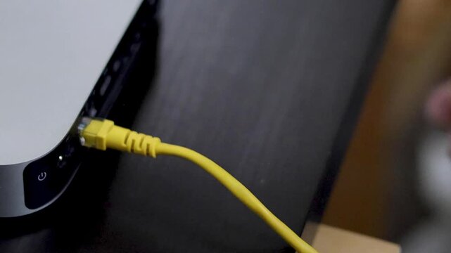Person plugging yellow ethernet cable into port of silver mini computer on wooden desk.