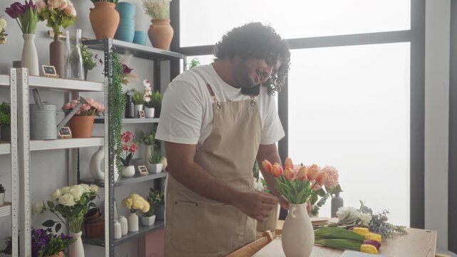 Man florist wearing apron holds tulip while arranging a vase at studio counter, speaking on smartphone at his ear near shelves and window; craft care serenity.