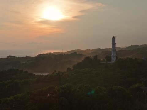 Aerial view of the white Mercusuar Baron lighthouse standing on a lush green hill overlooking the ocean during a golden sunset in Tanjungsari, Special Region of Yogyakarta, Indonesia.