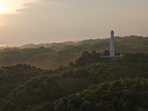 Aerial view of the white Baron Beach lighthouse standing on a lush green hill surrounded by misty tropical forests during sunset in Tanjungsari, Special Region of Yogyakarta, Indonesia.