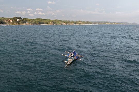 Aerial view of a traditional fishing boat with a fisherman on the blue sea near Baron Beach with lush green hills in the background in Tanjungsari, Special Region of Yogyakarta, Indonesia.