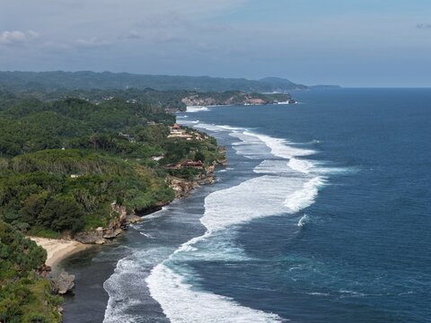 Aerial view of Baron Beach with white waves crashing against lush green cliffs and the blue ocean in Tanjungsari, Special Region of Yogyakarta, Indonesia.