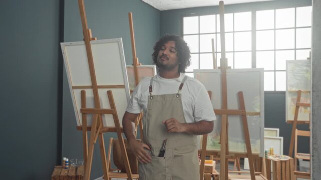 Young indian man painter in studio holds paintbrushes and gestures with raised hand toward canvas while wearing apron; focused creativity.