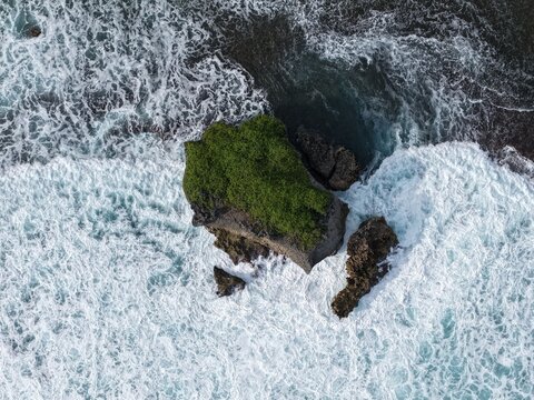 Aerial view of a small green islet surrounded by crashing white waves and turquoise water at Baron Beach Tanjungsari, Special Region of Yogyakarta, Indonesia.