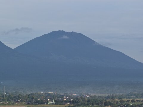 Aerial view of a massive volcanic mountain towering over a residential town with houses and lush trees under a hazy sky in Tanjungsari, Special Region of Yogyakarta, Indonesia.