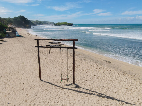 Aerial view of Baron Beach with a wooden swing on the sand, turquoise waves, and green cliffs in Tanjungsari, Special Region of Yogyakarta, Indonesia.
