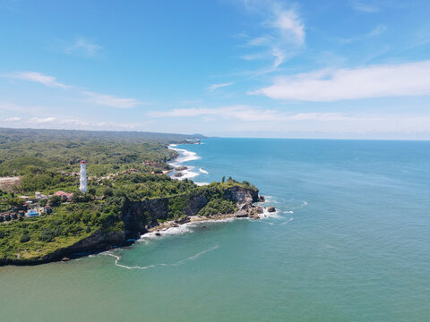 Aerial view of Baron Beach with its white lighthouse on the cliffs overlooking the turquoise sea and lush green hills Tanjungsari, Special Region of Yogyakarta, Indonesia.