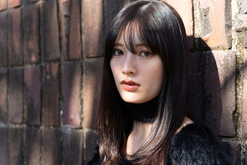 Fototapeta premium レンガの壁を背に、黒いファーニットを着て佇む若い日本人女性のポートレート