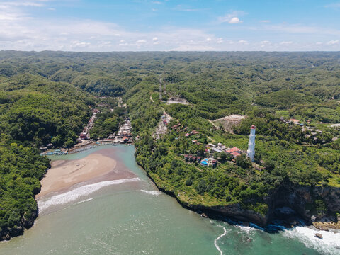 Aerial view of Baron Beach with its iconic white lighthouse, rugged cliffs, and turquoise waters surrounded by lush tropical hills Tanjungsari, Special Region of Yog, Indonesia.