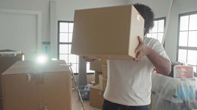 Man with arms crossed beside stacked cardboard moving boxes and bubble wrap, unpacking tools and plastic covered furniture visible in building; confidence.