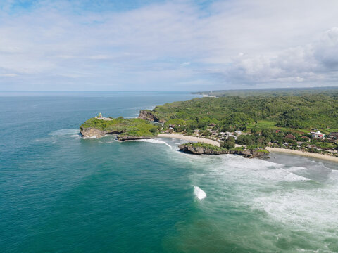Aerial view of Baron Beach featuring turquoise ocean waves crashing against rugged limestone cliffs and lush green hills in Tanjungsari, Special Region of Yogyakarta, Indonesia.