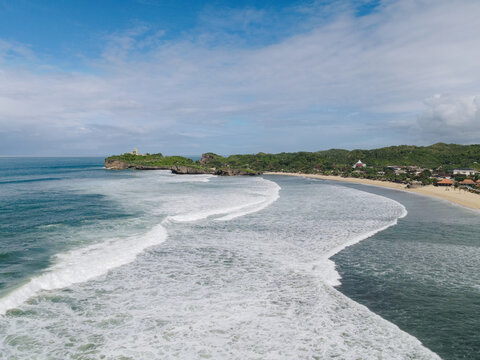 Aerial view of Baron Beach with crashing white waves along the sandy shoreline and lush green cliffs in Tanjungsari, Special Region of Yogyakarta, Indonesia.