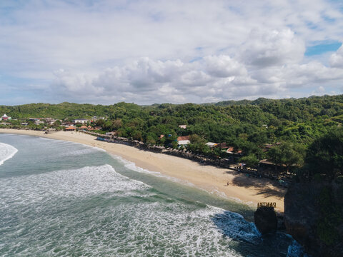 Aerial view of Baron Beach featuring the sandy shoreline, rolling ocean waves, and lush green hills under a cloudy sky in Tanjungsari, Special Region of Yog, Indonesia.