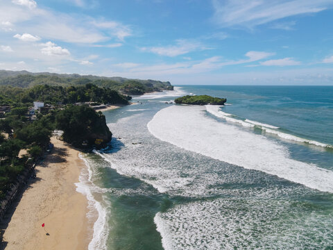 Aerial view of Baron Beach featuring golden sand, turquoise ocean waves crashing against the shore, and lush green cliffs under a blue sky in Tanjungsari, Special Region of Yogyakarta, Indonesia.