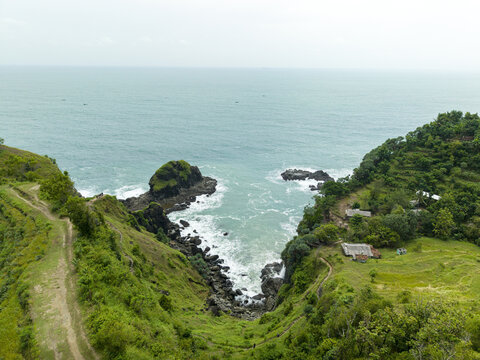 Aerial view of Banyutibo Water Fall cascading from lush green cliffs into the turquoise ocean with rocky shores in Girisubo, Special Region of Yogyakarta, Indonesia.
