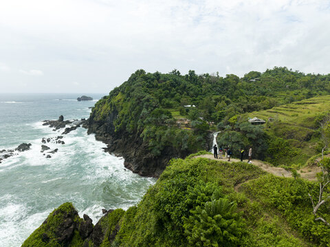 Aerial view of Banyutibo Water Fall cascading down rocky cliffs into the ocean with people standing on a grassy ridge Girisubo, Special Region of Yogyakarta, Indonesia.
