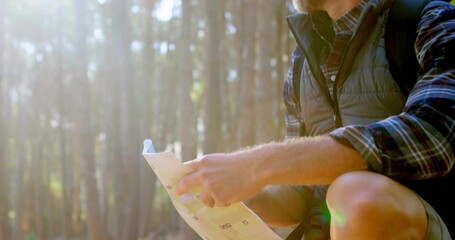 Naklejka premium Crouching hiker wearing plaid shirt and vest holding map in conifer forest, backpack, copy space
